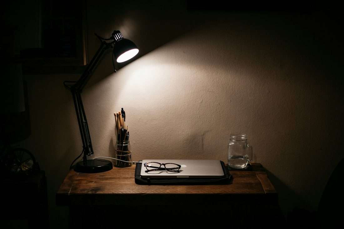 Dunkler Schreibtisch bei Nacht, die Lampe brennt, der Laptop ist zugeklappt, die Brille liegt darauf